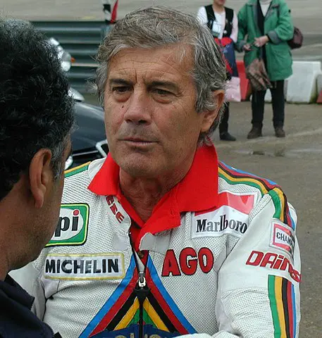 Giacomo Agostini seen in the photo wearing a red and white jacket and talking to the man who is partially seen. Agostini has white hair and this is well after his retirement from racing.