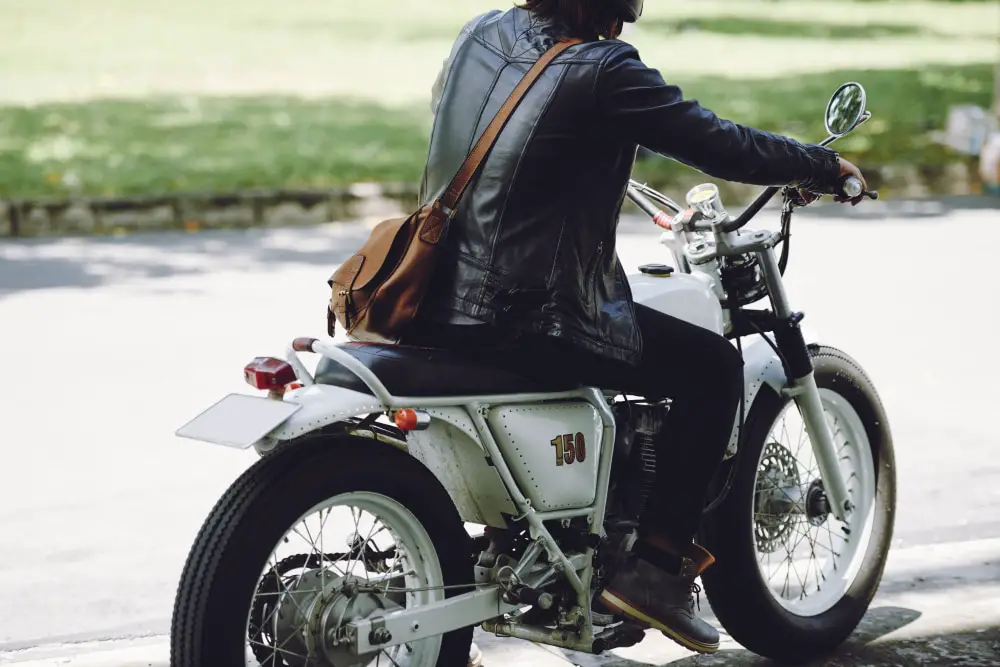 Motorcycle rider during the day sitting on his bike. The rider is wearing a helmet which can only be partially seen and is on a naked motorcycle white in color. We can only see the rider from the side and slightly from behind. The rider is wearing a black leather jacket.