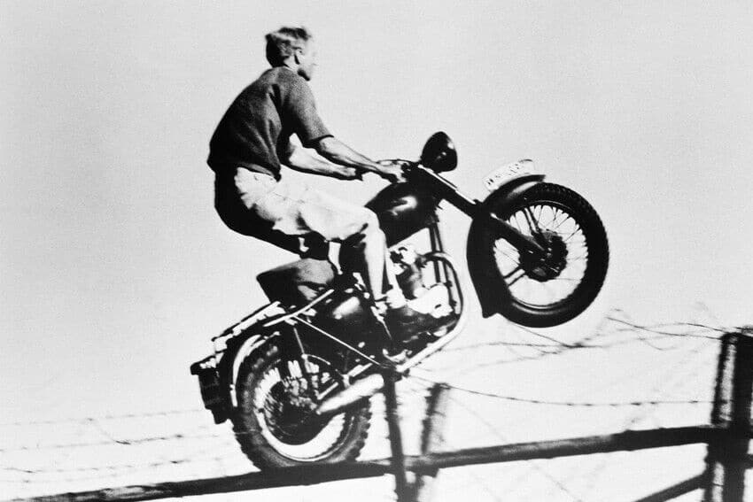 B & W photo of Steve McQueen on his motorcycle jumping over the fence in the movie the great escape.