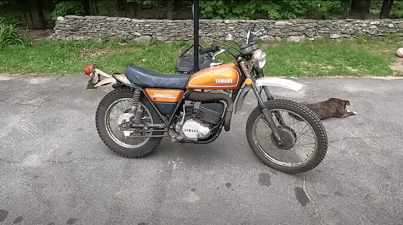 An old Yamaha motorcycle orange in color. There is black dog in the back of the motorcycle resting on the tarmac with his face turned away. The motorcycle seem to be a 300 or 400 cc engine capacity bike. The setting looks like rural.